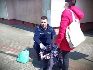 policjant kuca przy kobiecie siedzącej na chodniku, obok nich stoi druga kobieta