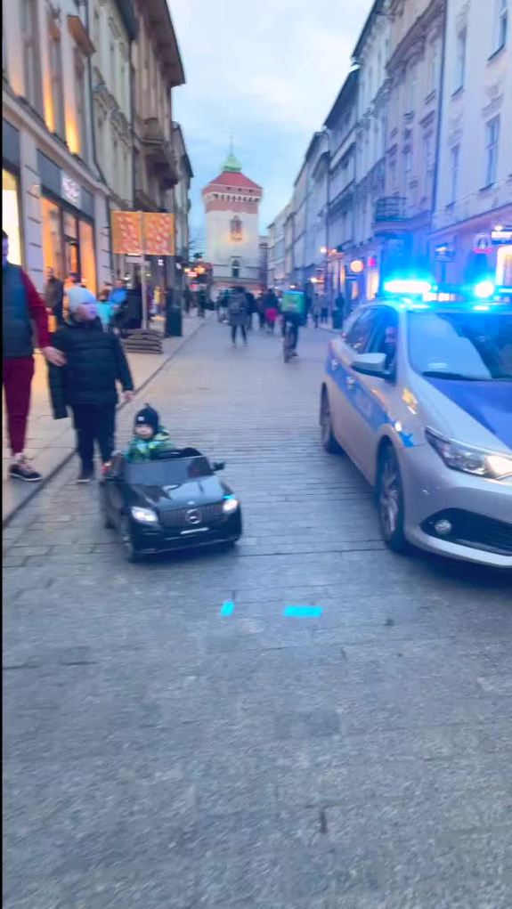 Dziecko w miniaturowym samochodziku obok policyjnego radiowozu z włączonymi sygnałami na wąskiej uliczce starówki.
