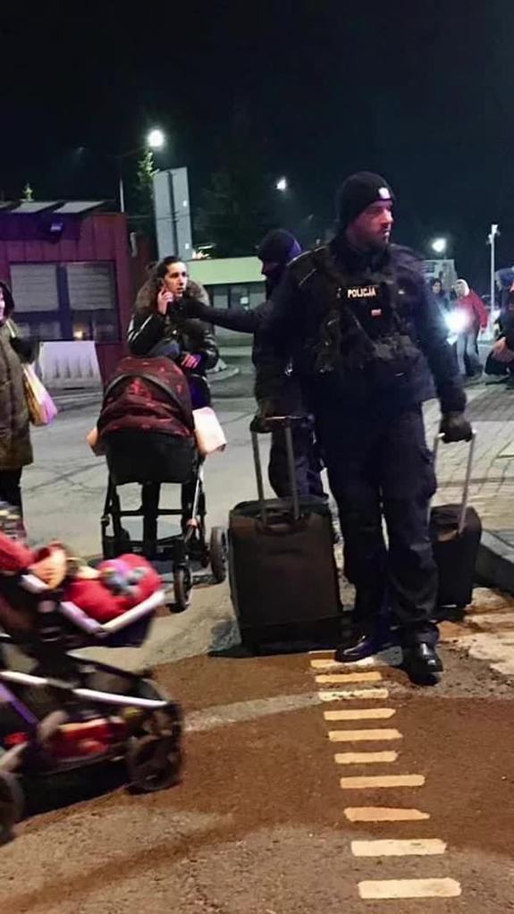 Policjant trzyma dwie walizki na kółkach, obok niego młoda kobieta z wózkiem dziecięcym rozmawia przez telefon.