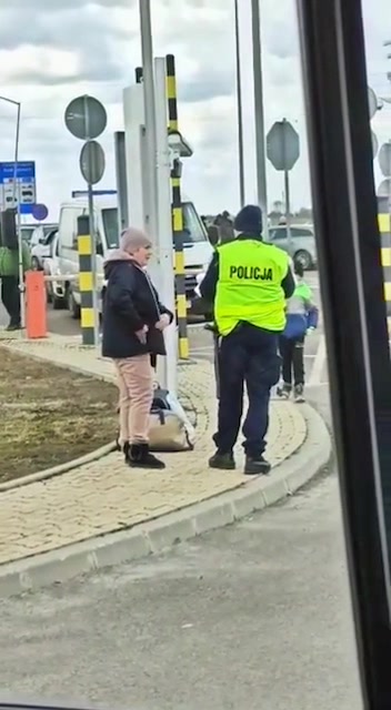 Policjant w żółtej kamizelce z napisem POLICJA na plecach rozmawia z kobietą.