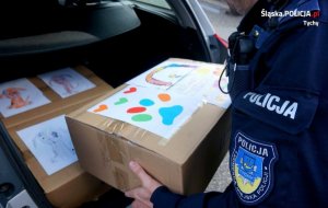 Na zdjęciu widoczna ręka policjanta w mundurze, który wkłada do bagażnika karton, na którym są rysunki dzieci.&quot;&gt;