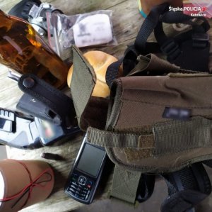 zdjęcie kolorowe stołu, na którym znajduje się butelka po piwie, torba typu nerka, telefon komórkowy, materiały pirotechniczne, paralizator, bułka