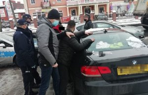 Oszuści podczas zatrzymania przez policję. Mężczyźni oparci są o samochód marki BMW. Obok policjanci umundurowani oraz po cywilnemu w tle radiowóz