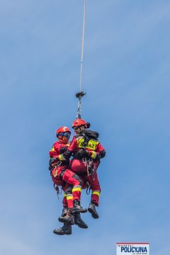 Zbliżenie na dwóch strażaków zwisających na linie w powietrzu.