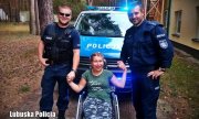dwaj umundurowani policjanci stoją przed radiowozem i trzymają za dłonie dziewczynę na wózku
