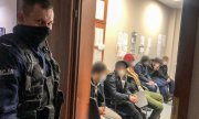 policjant na korytarzu w komendzie, a w pomieszczeniu mężczyźni siedzący na krzesłach - twarze mężczyzn zamazane graficznie