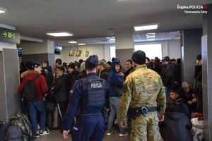 policjant w niebieskim mundurze i funkcjonariusz straży granicznej obserwujący liczną grupę pasażerów czekających w kolejce, w terminalu lotniska