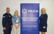 Na środku baner z napisem Komenda Powiatowa Policji w Mońkach. Z prawej strony baneru stoi kobieta w sukience, z lewej kobieta w spodniach  a obok niej umundurowany policjant