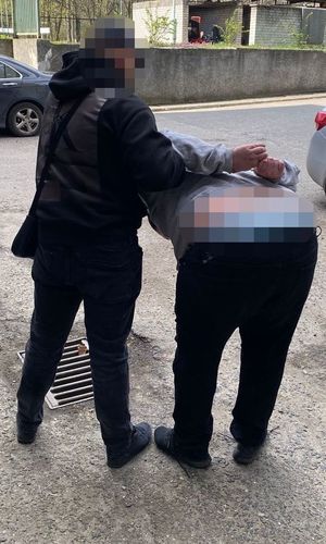 Zdjęcie przedstawia policjanta ubranego w cywile rzeczy, który prowadzi po swojej prawej stronie zatrzymanego mężczyznę. Mężczyzna ten jest zgięty w pół i pochylony do przodu. Na rękach trzymanych z tyłu ma założone kajdanki.
