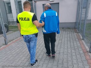 policjant w żółtej kamizelce z napisem policja prowadzi zatrzymanego mężczyznę w niebieskiej bluzie