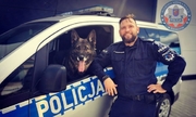 policjant stoi przy radiowozie, przez okno radiowozu wystaje głowa psa policyjnego