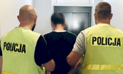 Odwróceni tyłem dwaj policjanci w żółtych kamizelkach z napisem policja z zatrzymanym mężczyzną zakutym w kajdanki