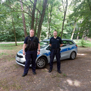 dwaj policjanci stoją przed radiowozem policyjnym, w tle drzewa