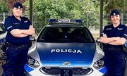 Dwie umundurowane policjantki stoją po bokach oznakowanego radiowozu