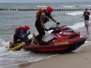 ratownik na brzegu morza na skuterze wodnym, z tyłu drugi ratownik z tyłu udziela pomocy osobie