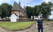 na pierwszym planie widać umundurowanego policjanta, który stoi przed kościołem w miejscowości Blizne, będący jednocześnie zabytkiem wpisanym na światową listę UNESCO. W tle zdjęcia widoczna zabudowa sakralna z otaczającym kościół murem oraz sam budynek kościelny
