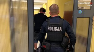 Policjanci prowadzą podejrzanego mężczyznę korytarzem, przechodzą przez drzwi.