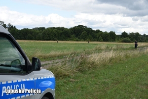 radiowóz i policjanci na poszukiwaniach na terenie trawiastym, daleko w tle drzewa i krzewy