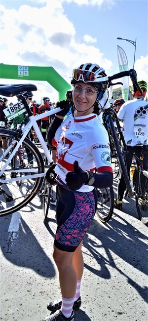 Kobieta w stroju sportowym jako zawodniczka, trzyma na ramieniu rower, stojąc  przed metą