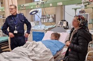 dziecko leży na łóżku w szpitalu, obok niego stoi kobieta i policjant