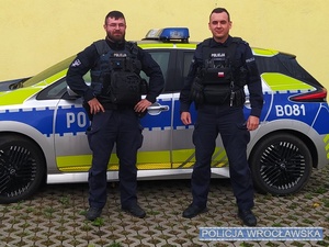 Dwaj umundurowani policjanci stojący na tle oznakowanego radiowozu Policji