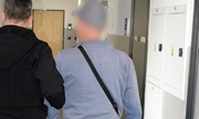 po lewej stronie policjant pionu kryminalnego trzymający pod rękę zatrzymanego ubranego w dżinsy, szarą bluzę i szarą czapeczkę z daszkiem. Przepasaną ma również czarną małą torbę
