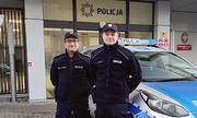 Dwóch policjantów stojących przy radiowozie, w tle budynek komisariatu