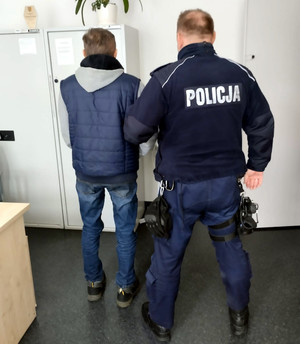 Policjant w kamizelce z napisem Policja na plecach stoi z zatrzymanym w pomieszczeniu. Widok z tyłu