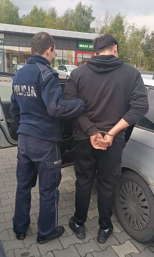 policjant stoi z zatrzymanym mężczyzną. Obaj stoją przy pojeździe osobowym, w tle parking i sklep