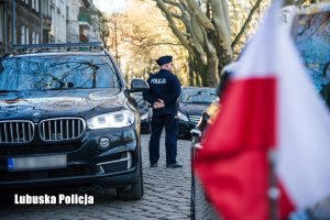 Policjant stojący na drodze, a obok niego pojazd osobowy. Na pierwszym planie flaga Polski.
