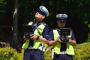 Dwaj policjanci ruchu drogowego, umundurowani i w odblaskowych kamizelkach, trzymają w rękach panel sterowania dronem.