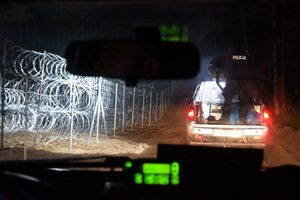 Zdjęcie wykonane w nocy z wnętrza policyjnego radiowozu. Po lewej zabezpieczenia graniczne i drut kolczasty. Po prawej policyjny radiowóz terenowy z otwartą częścią z tyłu, na której stoi policjant, kierujący strumieniem światła z reflektora.