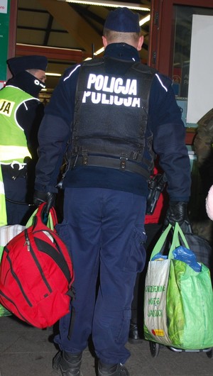 Zdjęcie kolorowe wykonane w porze nocnej przedstawia dworzec PKP w Przemyślu- na zdjęciu widoczny policjant umundurowany który pomaga uchodźcom w przenoszeniu bagaży