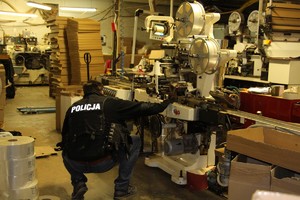 Policjant ubrany w czarną kamizelkę z białym napisem &quot;policja&quot;  klęczy tyłem  przy maszynie wykorzystywanej do produkcji podrabianych papierosów, w tle pomieszczenia nielegalnej fabryki z kartonami i innymi maszynami
