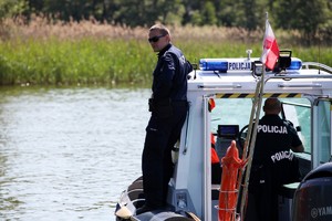 Policyjna motorówka płynie po jeziorze,  a na jej boku stoi policjant w mundurze i okularach słonecznych