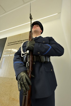Zdjęcie. Umundurowany policjant pełniący wartę honorowa przed tablicą