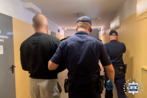 Policjanci prowadzą zatrzymanego mężczyznę idąc korytarzem.