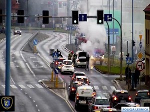 Widok z kamery miejskiej na zatłoczoną ulicę i policjanta i strażaków gaszących płonący samochód