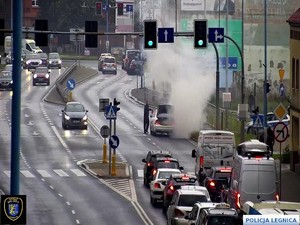 Widok z kamery miejskiej na zatłoczoną ulicę i policjanta gaszącego pożar płonącego na drodze auta