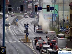 Widok z kamery miejskiej na zatłoczoną ulicę i policjanta gaszącego płonący samochód