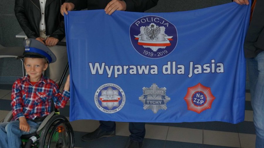chłopiec na wózku inwalidzkim w policyjnej czapce trzyma flagę z napisem "Wyprawa dla Jasia" 