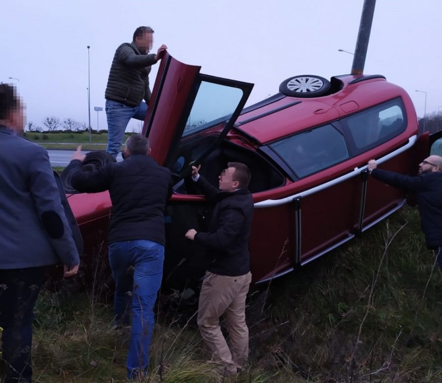 Na fotografii widać leżący na boku w rowie samochód osobowy. Na samochodzie stoi mężczyzna który trzyma otwarte drzwi  od strony pasażera. Kolejni mężczyźni trzymają samochód, aby nie przewrócił się na dach