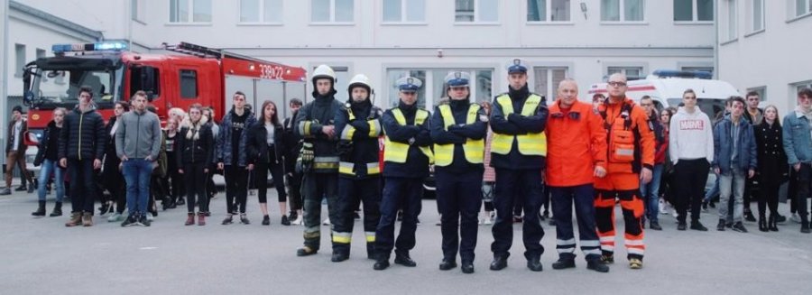 słuzby mundurowe stoją przed budynkiem za nimi stoją osoby którym pomagają na co dzień 