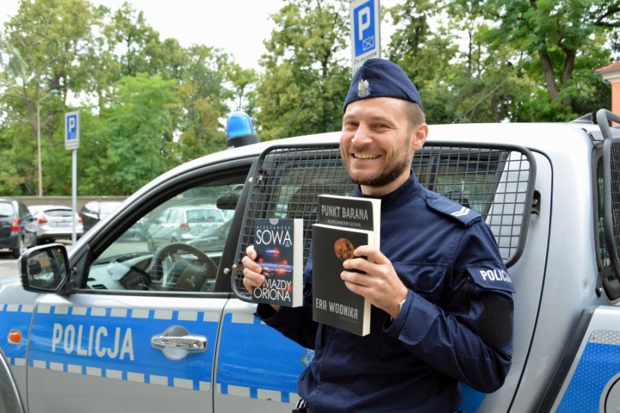 Sierż. sztab. Aleksander Sowa stoi przed radiowozem i trzyma swoje książki