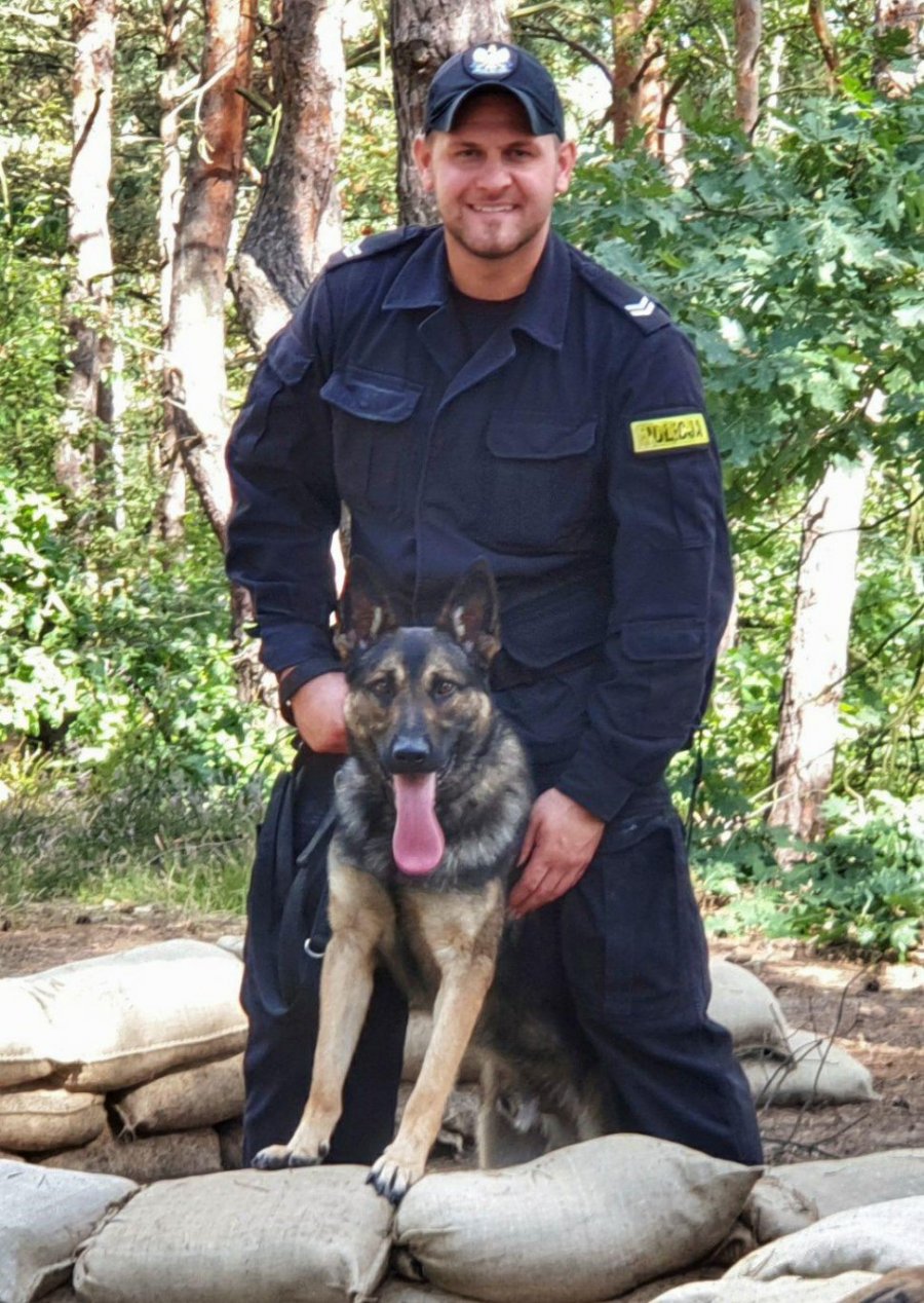 Na zdjęciu widać st. sierż. Kamila Kubicę, który trzyma na smyczy psa patrolowo-tropiącego. Policjant i pies znajdują się w okopie z workami piachu