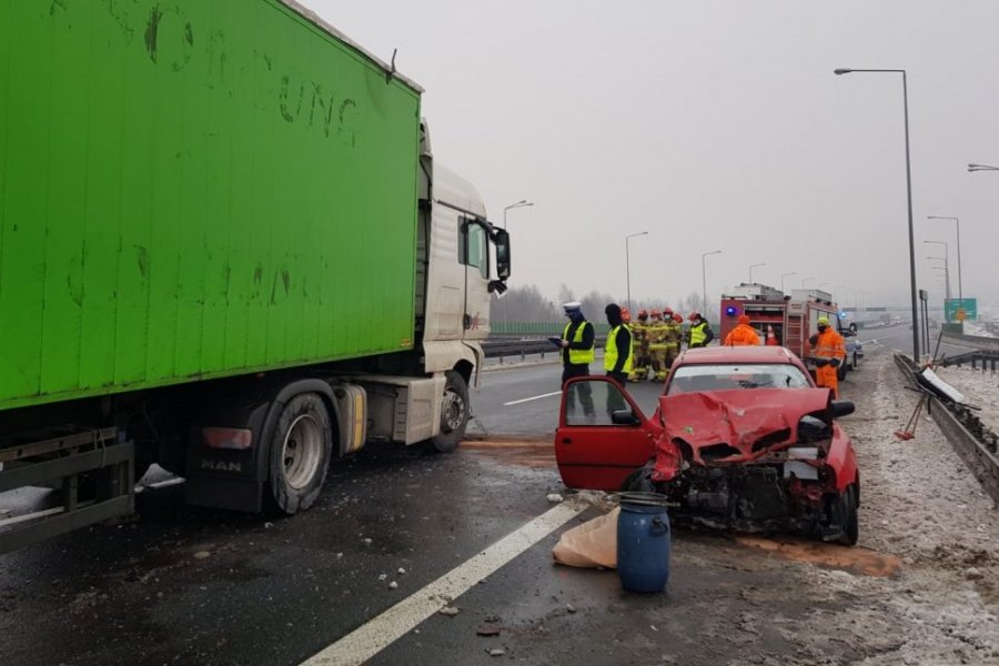 Na pasie jezdni stoi TIR z naczepą, z prawej strony czerwony samochód osobowy z rozbitym przodem. Pomiędzy nimi stoi beczka z odczynnikiem do czyszczenia jezdni z płynów eksploatacyjnych pojazdów.