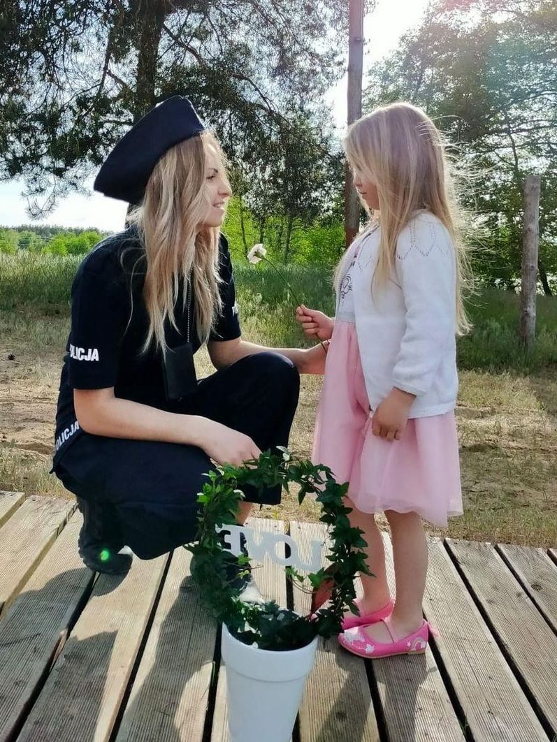 mała dziewczynka daje kwiatka swojej mamie - policjantce w mundurze. Przed nimi w białej doniczce znaduje się zielone kółko uformowane z rosłiny i biały napis LOVE
