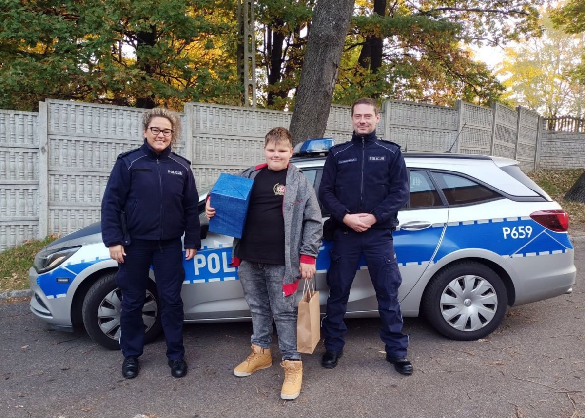 Od lewej stoją: umundurowana policjantka, obok niej chłopiec, który pod jedną pachą trzyma prezent, w drugiej ręce papierową torebkę, dalej umundurowany policjant. Za nimi stoi policyjny radiowóz