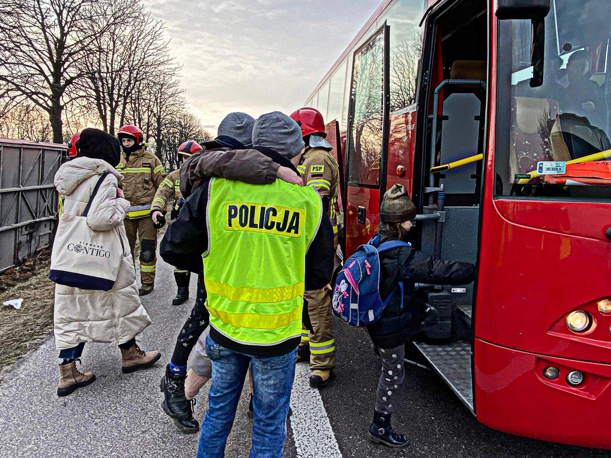 Policjant trzyma na rękach dziecko. Poszkodowani w wypadku wsiadają do zastępczego autokaru