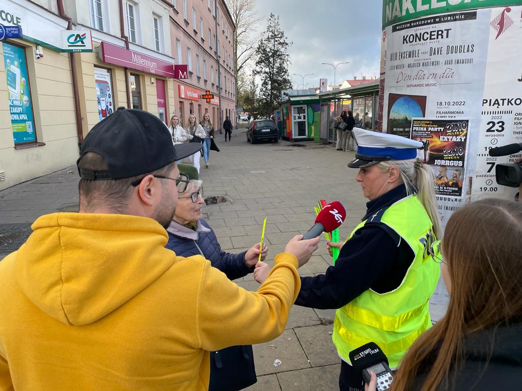 Umundurowana policjantka w kamizelce odblaskowej wręcza kobiecie element odbaskowy. Z boku stoi dziennikarz radia Zet, który trzyma przed policjantka redakcyjny mikrofon